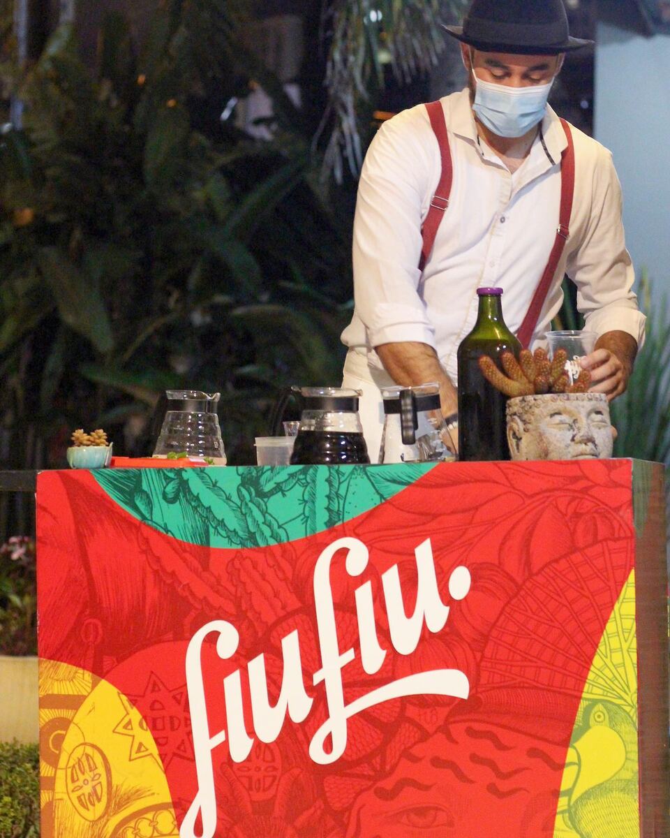 Bartender de chapéu e suspensórios preparando um drink atrás de um balcão coberto com tecido colorido