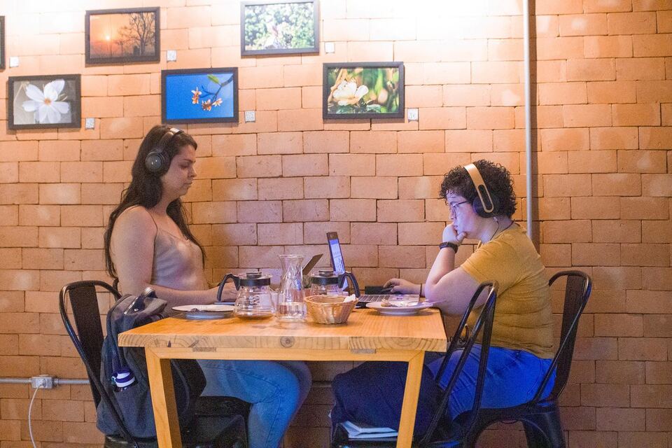 Duas mulheres de fone de ouvido gravando numa mesa do salão, com fotos emolduradas na parede de tijolos