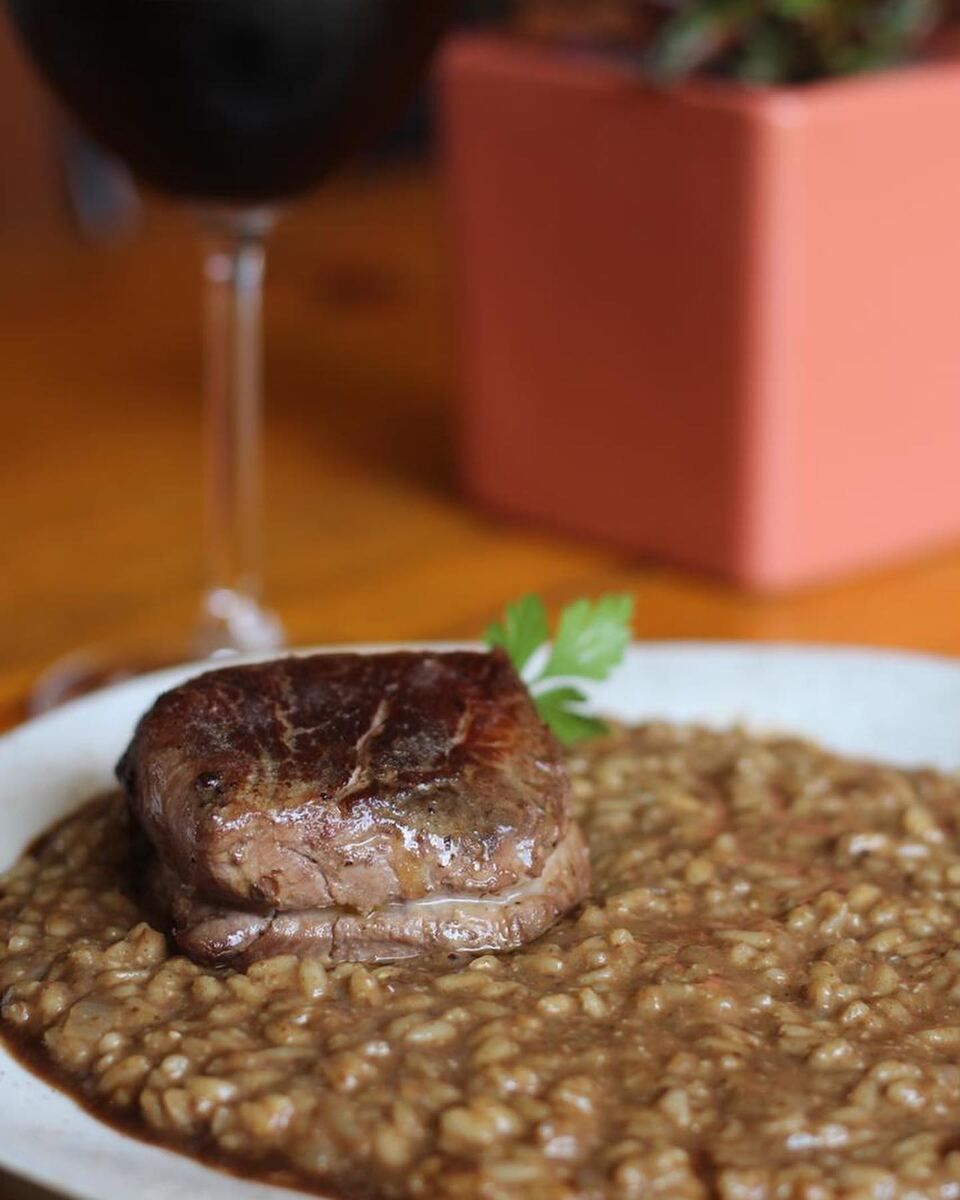 Medalhão de carne sobre risoto escuro, com uma taça de vinho tinto ao fundo