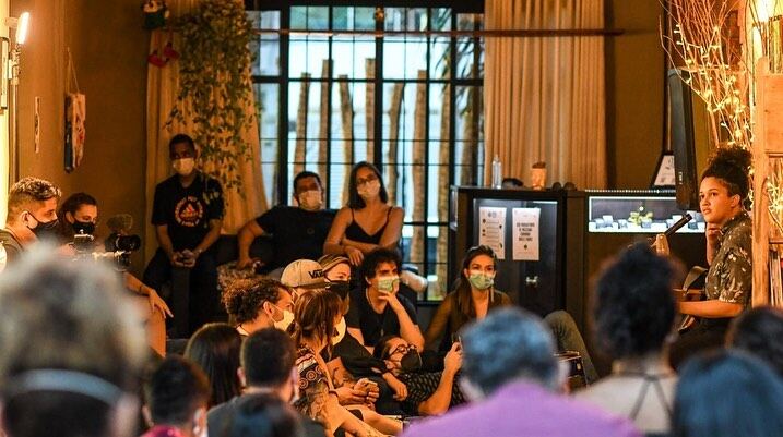 Público reunido no salão à noite, ouvindo uma artista que fala em pé, com luz quente