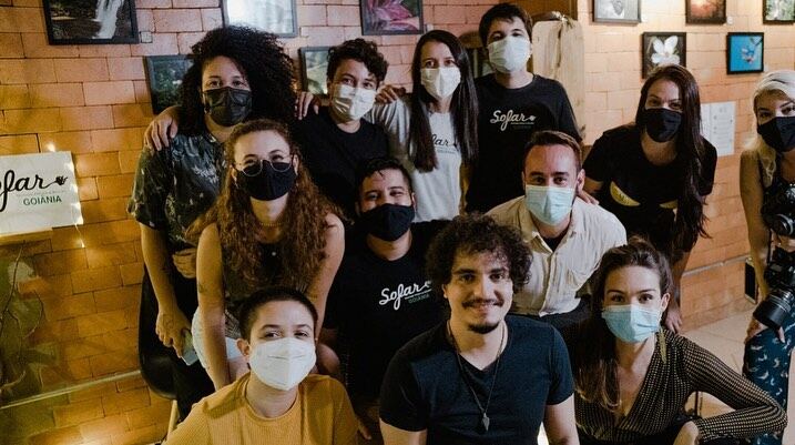 Grupo grande de público e artistas do Sofar Sounds reunidos para a foto, de máscara, na parede de tijolos da casa