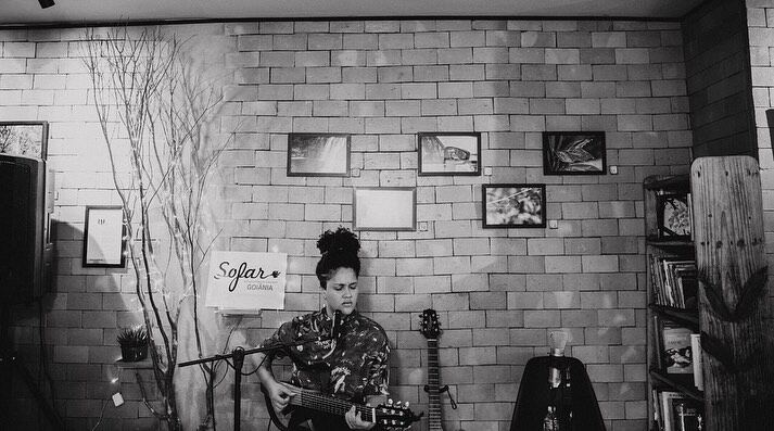 Foto em preto e branco de uma cantora com violão num show intimista do Sofar, com quadros na parede ao fundo